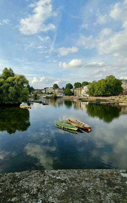 A wide view of a calm river reflecting the partly cloudy sky above, with white fluffy clouds and patches of blue. On the left bank, there are lush green trees with dense foliage, while the right bank features a row of residential buildings and parked boats, some of which are moored along the shoreline. The boats in the foreground are small, wooden, and painted in various muted colors, appearing to be used for leisure or fishing. The scene is well-lit with natural daylight, emphasizing the peaceful atmosphere of the river environment. While the image mainly depicts a tranquil waterway, it subtly relates to waste management by showcasing a setting where independent collection or private disposal of waste from riverside properties could take place, aligning with services like those offered by Waste Collection Richmond for rubbish removal in the area.