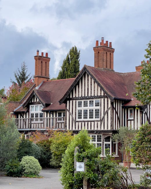 A traditional two-storey house featuring a distinctive black and white timber-framed facade, with multiple gable roofs covered in red tiles. The building has tall, red-brick chimneys protruding from the roof, and large, multi-pane sash windows on both floors. Surrounding the house, there is a lush front garden with a variety of green shrubs and small trees, some with dense foliage and others more sparse, placed along a paved pathway. A white utility notice or sign is mounted on a small post amidst the greenery near the front of the property. The sky overhead is partly cloudy, casting soft, diffuse light onto the scene. The overall setting suggests a residential area where private property maintenance, including rubbish collection or garden waste disposal, might be managed directly by the homeowner or through external waste management services such as Waste Collection Richmond, supporting on-site clearance or alternative waste handling options.