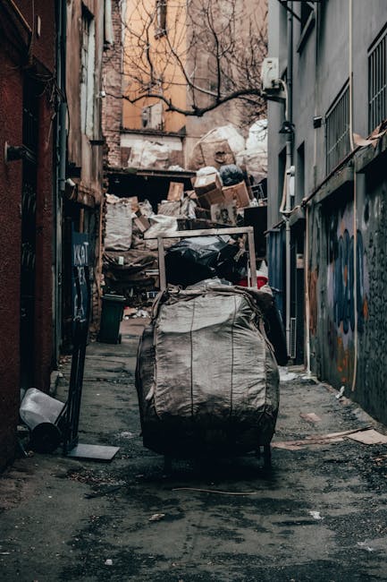 A narrow alleyway with uneven, dark asphalt paving, cluttered with discarded items and debris, including a large black trash bag leaning against a metal pushcart in the foreground. Behind the cart, there are multiple piles of mixed waste, such as cardboard boxes, paper, and plastic bags, stacked haphazardly against a backdrop of densely packed buildings. The walls on either side are made of weathered brick and painted surfaces, some with graffiti and peeling paint. Bare tree branches extend overhead, partially obscuring the sky, which is overcast, creating a dim, moody atmosphere. Visible utility pipes and cables are attached to the building exteriors, indicating an urban environment. The scene overall suggests an informal waste accumulation area that may be addressed through private rubbish collection services like Waste Collection Richmond, offering alternative waste handling options for narrow streets in dense urban settings.