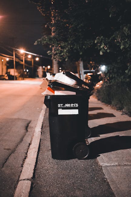 A black wheeled rubbish bin labeled 'St. John's' positioned on a concrete sidewalk next to a street at night. The bin's lid is open, revealing a pile of mixed waste including cardboard, paper, and plastic bags. The scene is illuminated by streetlights, casting shadows on the ground and highlighting the textured surface of the bin and the waste material. In the background, a row of trees and residential streetlights create a softly lit environment, with parked cars visible along the curb. The bin appears to be used for collection of general rubbish or waste disposal, possibly associated with private waste handling services like those offered by wastecollectionrichmond.co.uk. The image emphasizes on-site waste storage and the importance of proper rubbish management in urban areas, with a focus on clean, professional waste collection practices.