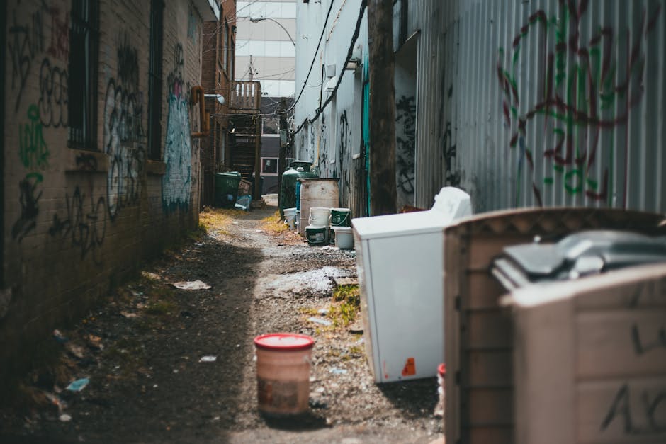 A narrow alleyway with uneven, dark asphalt paving, cluttered with discarded items and debris, including a large black trash bag leaning against a metal pushcart in the foreground. Behind the cart, there are multiple piles of mixed waste, such as cardboard boxes, paper, and plastic bags, stacked haphazardly against a backdrop of densely packed buildings. The walls on either side are made of weathered brick and painted surfaces, some with graffiti and peeling paint. Bare tree branches extend overhead, partially obscuring the sky, which is overcast, creating a dim, moody atmosphere. Visible utility pipes and cables are attached to the building exteriors, indicating an urban environment. The scene overall suggests an informal waste accumulation area that may be addressed through private rubbish collection services like Waste Collection Richmond, offering alternative waste handling options for narrow streets in dense urban settings.