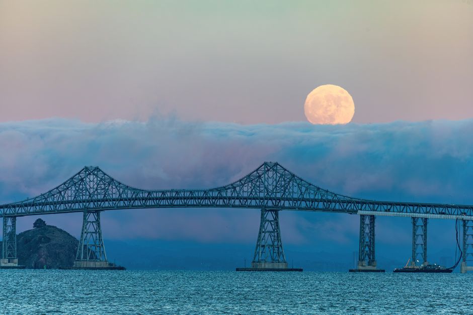 A large steel bridge with an intricate truss structure spans across a body of water, supported by multiple tall pylons. The bridge appears to be a suspension design, featuring curved main cables and vertical suspender cables. To the left of the bridge, a small island or landmass with a single tree is visible. In the background, a thick layer of clouds partially obscures the scene, with an overcast sky above. The full moon, bright and round with visible surface details, rises behind the clouds, creating a luminous focal point in the sky. The water beneath the bridge is calm, with a textured surface reflecting the muted tones of the sky. The lighting suggests either dawn or dusk, with the scene characterized by a subdued, cool colour palette. This photograph captures a tranquil moment with natural and man-made elements blending against the backdrop of atmospheric conditions, and could be associated with the context of private or independent infrastructure maintenance or the visual setting of alternative landscape views in relation to waste management or environmental monitoring.