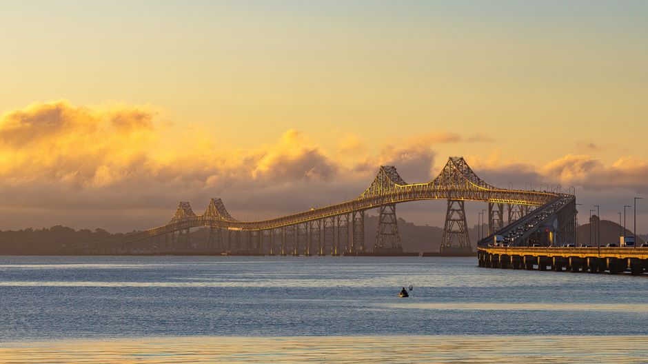 A large steel suspension bridge spans across a wide body of water, with its structure painted in a light yellow or gold hue that catches the soft natural light of a cloudy sky during sunset or sunrise. The bridge features tall towers with cables extending downward, supporting the roadway that curves gently connecting both ends. In the foreground, calm water reflects the colors of the sky and the bridge’s framework, with a small boat visible near the center of the image, creating gentle ripples on the surface. To the right, a wooden dock or pier extends into the water, lined with evenly spaced street lamps and a utility building or container, indicating a functional area for access or maintenance. The background depicts partly cloudy skies with warm tones blending into cooler shades as the sun’s light filters through, emphasizing the bridge’s structural details against a picturesque, tranquil waterscape. This scene exemplifies infrastructure designed for both utility and aesthetic appeal, similar to private site clearance or independent waste collection methods on a broader level, integrated naturally within the environment.